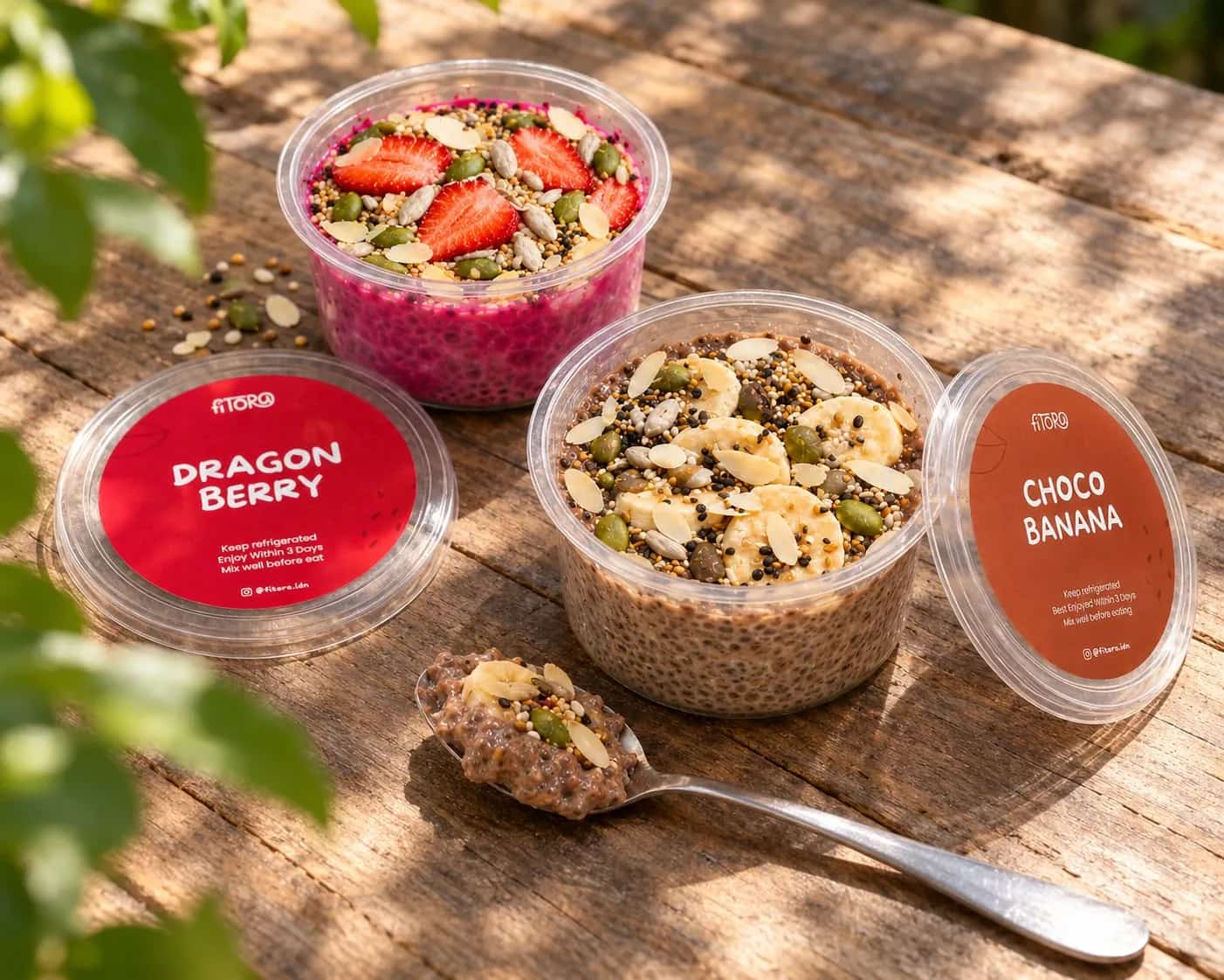 Chia Pudding Dragon Berry dengan stroberi dan Choco Banana dengan pisang, terbuka di meja kayu, tutupnya di samping, sesendok Choco Banana di depan