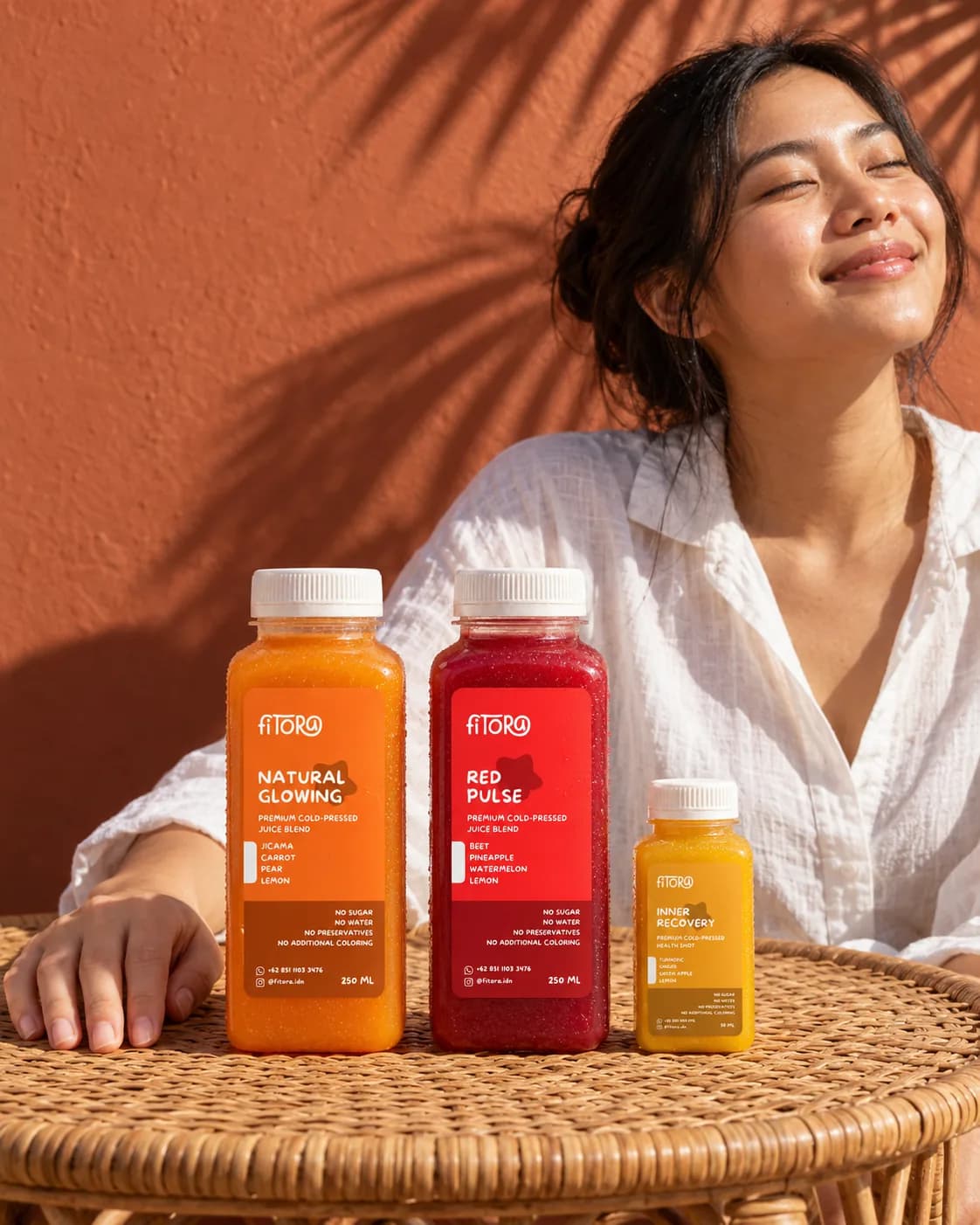 Perempuan berkemeja linen putih duduk di balik meja rotan, memejamkan mata di bawah matahari, dengan Natural Glowing, Red Pulse, dan shot Inner Recovery berjajar di meja.